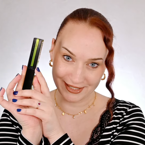 Frau zeigt lächelnd eine Mascara-Verpackung von Lethal Cosmetics in die Kamera; neutrale Studio-Umgebung, gestreiftes Oberteil, blaue Nägel.