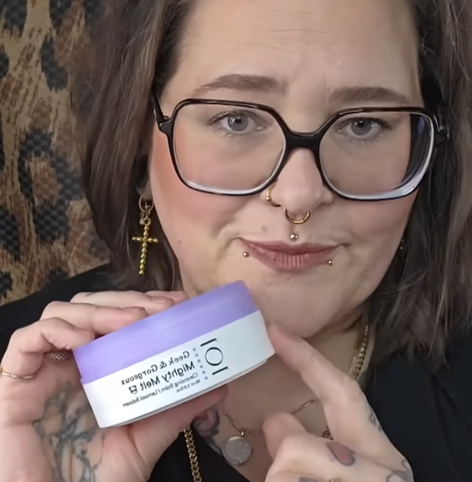 Frau mit Brille zeigt eine runde, lilafarbene Geek & Gorgeous Mighty Melt Reinigungspflege und deutet darauf; Hintergrund in Leopardoptik.