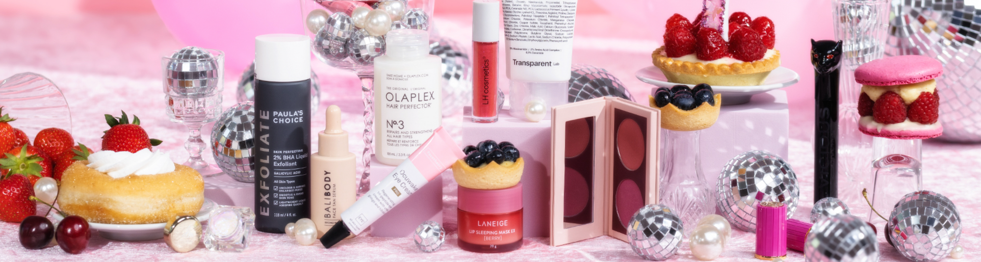 Verschiedene Schönheitsprodukte und Desserts auf einer rosa Fläche mit Discokugeln, Perlen und Beeren als Dekoration.