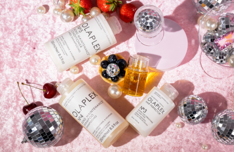 Olaplex-Haarpflegeprodukte auf rosafarbenem Stoff mit Perlen, Früchten und Mini-Diskokugeln verstreut.