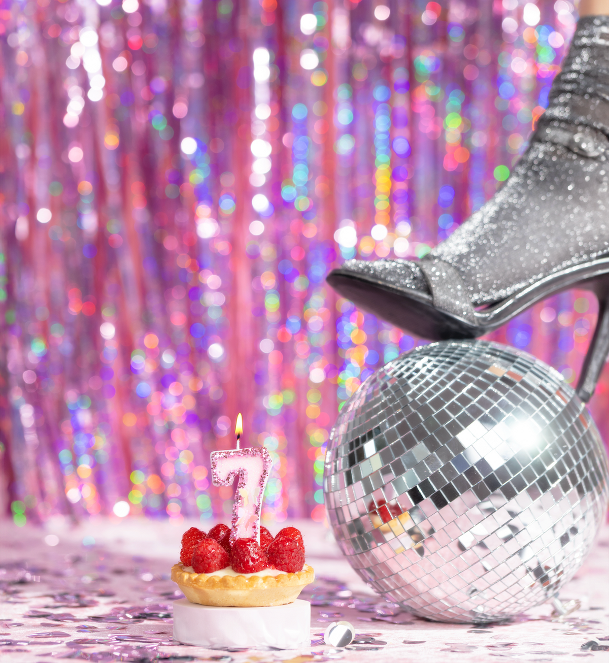 Eine brennende Kerze auf einer Torte, daneben eine Discokugel und ein glitzernder Schuh, vor einem glitzernden rosa Hintergrund.