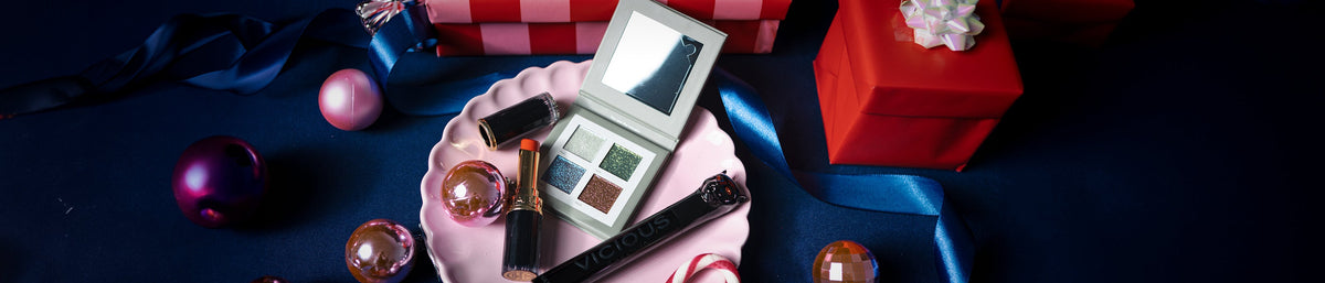 Auf einer dunklen Fläche steht ein Tablett mit Make-up-Artikeln und Schmuckstücken neben verpackten Geschenken und einem blauen Band.