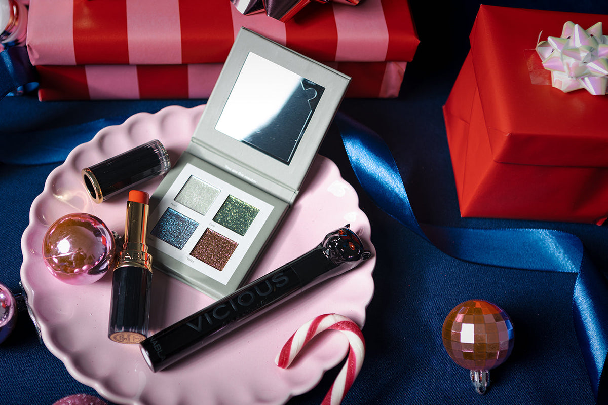 Make-up-Set mit Lidschattenpalette, Lippenstift und Mascara auf einem rosa Teller, umgeben von Geschenken, Schleifen und Dekorationsartikeln.