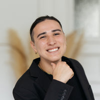 Eine Person mit dunklem Haar in einem schwarzen Blazer lächelt in die Kamera, wobei eine Hand unter dem Kinn ruht; die Aufnahme wurde in einem Innenraum gemacht.