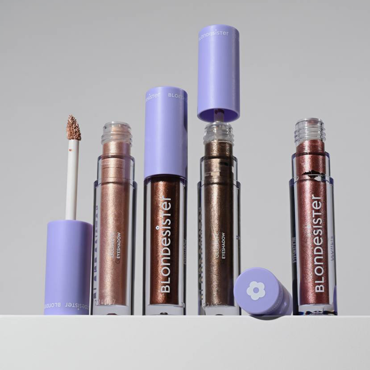 Drei GLOWEYE-Lidschatten-Tuben von BLONDESISTER mit violetten Verschlusskappen, von denen eine geöffnet ist, auf einer weißen Fläche vor grauem Hintergrund.