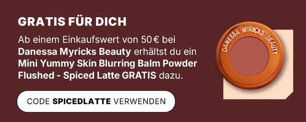 Werbegrafik mit dunkelrotem Hintergrund: Text informiert über eine Gratisaktion ab 50 € Einkaufswert bei Danessa Myricks Beauty. Abgebildet ist der Mini Yummy Skin Blurring Balm Powder Flushed in der Farbe Spiced Latte, in einer runden Verpackung mit kupferfarbenem Rand.