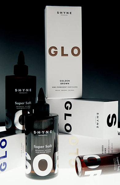 Flaschen und Schachteln von SHYNE GLO-Haarglanzprodukten, angeordnet auf einer reflektierenden Oberfläche mit dunklem Hintergrund.