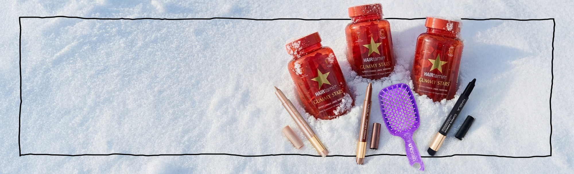 Drei HAIRtamin Gummy Stars im Schnee, umgeben von NABLA Cupid’s Arrow Stiften und einer lilafarbenen UNBrush-Haarbürste.