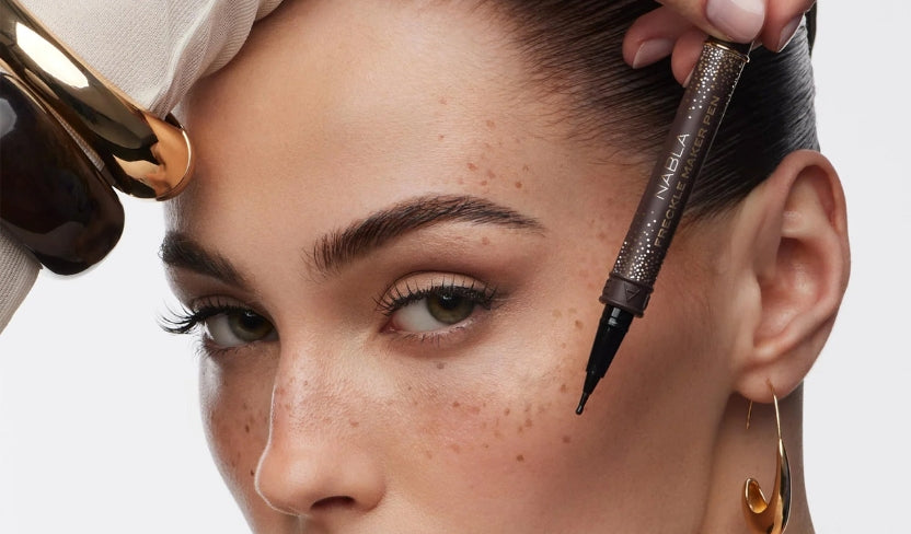 Eine Frau hält sich einen braunen Make-up-Stift an die Wange und präsentiert dabei ein natürliches Make-up sowie goldene Accessoires.
