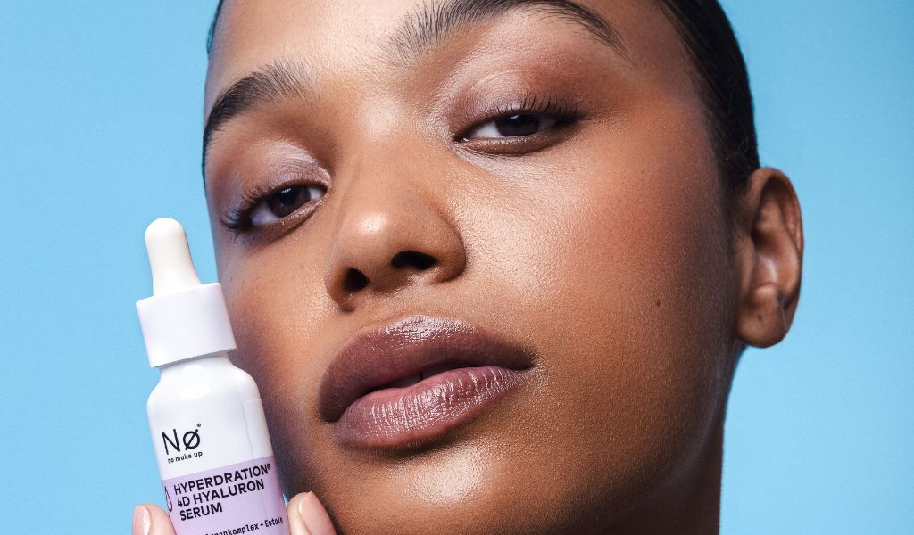 Frau mit gepflegter Haut hält ein Hyaluron-Serum von No Make Up in die Kamera, Nahaufnahme vor blauem Hintergrund.