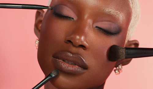 Frau mit geschlossenen Augen wird mit Make-up-Pinseln an Augen, Wange und Lippen geschminkt. Sanfter Look mit rosa Lidschatten.