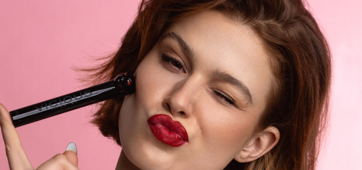 Eine junge Frau mit rotem Lippenstift zwinkert und drückt eine Mascara an ihre Wange vor einem rosa Hintergrund.