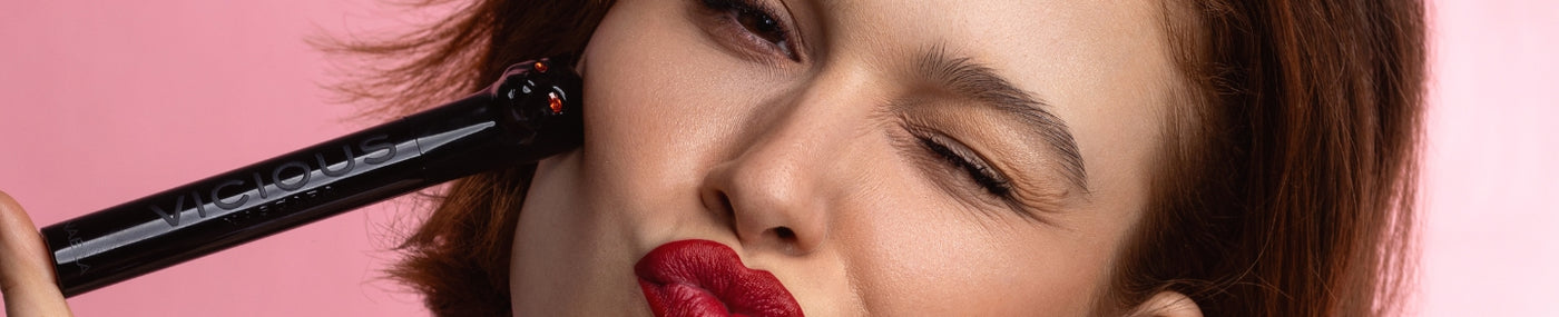 Eine Frau zwinkert mit rotem Lippenstift und hält eine Mascara an ihre Wange, vor einem rosa Hintergrund.