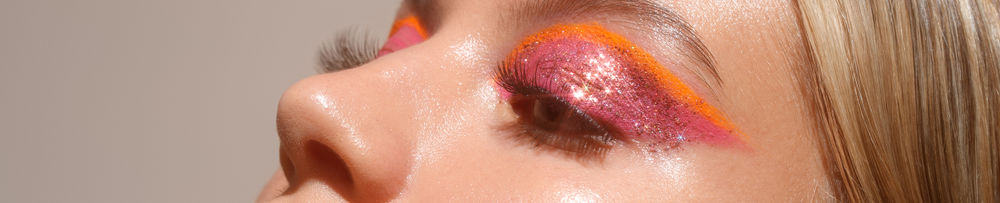 Detailaufnahme eines geschlossenen Auges mit glitzerndem pink-orangenem Lidschatten, langen Wimpern und glattem Teint.